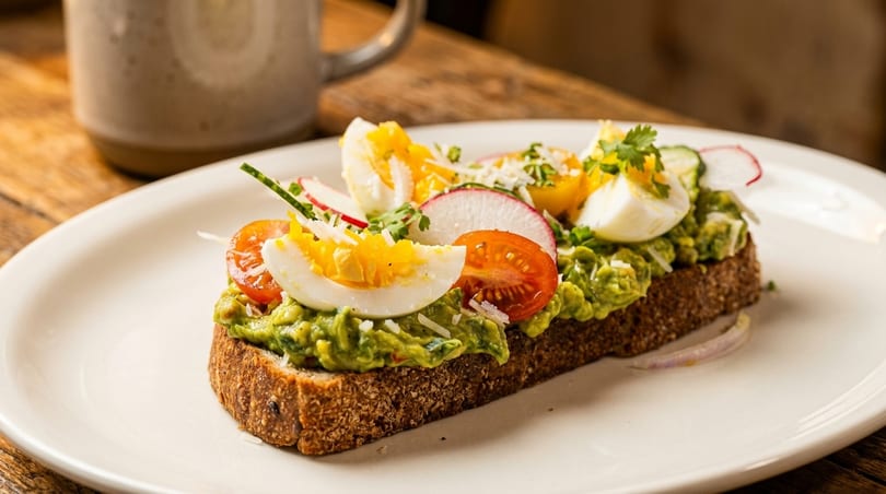 Avocado toast with sliced eggs, tomatoes, radishes, and herbs on a white plate.