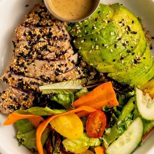 Seared tuna with sesame seeds, sliced avocado, mixed greens, and a side of dressing in a white bowl.