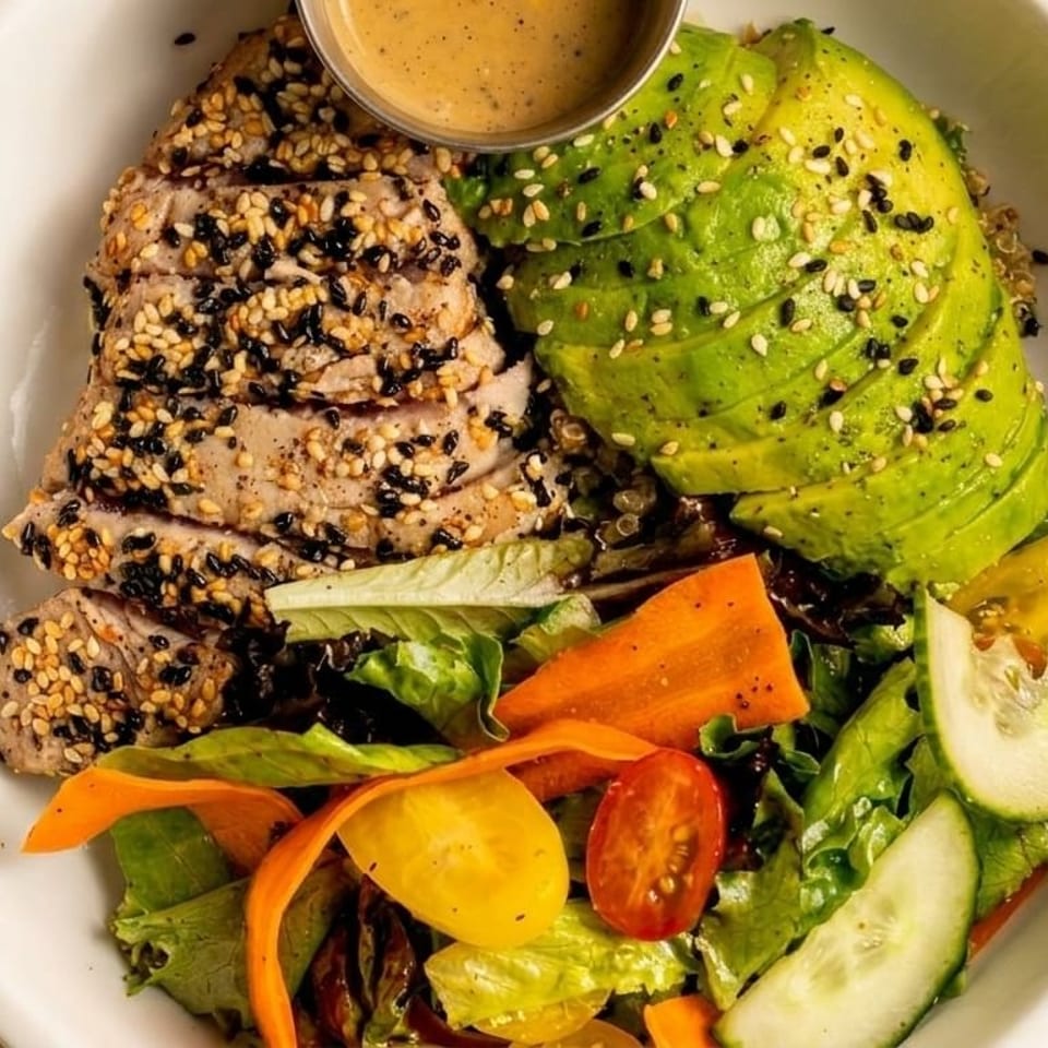 Seared tuna with sesame seeds, sliced avocado, mixed greens, and a side of dressing in a white bowl.